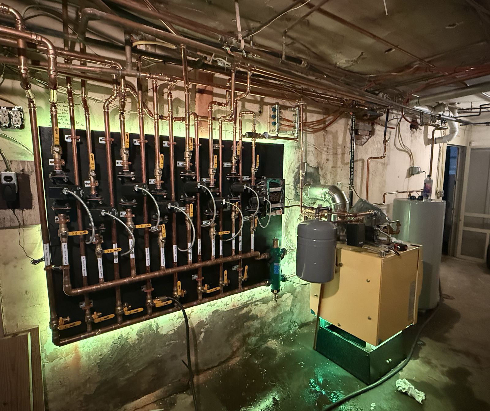 Basement boiler room with a copper multi-zone piping manifold and valves on a black board, next to a boiler, expansion tank and water heater
