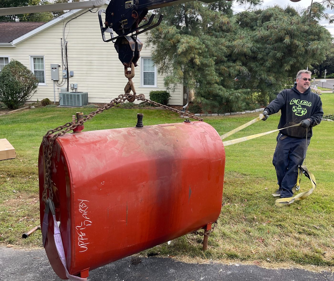 C&L technician guiding an old red heating oil tank as a truck crane lifts it from a yard