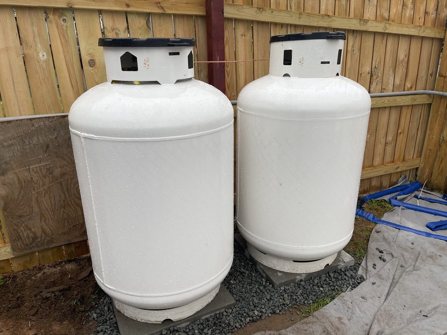 Two white residential propane tanks on a gravel pad beside a wooden fence
