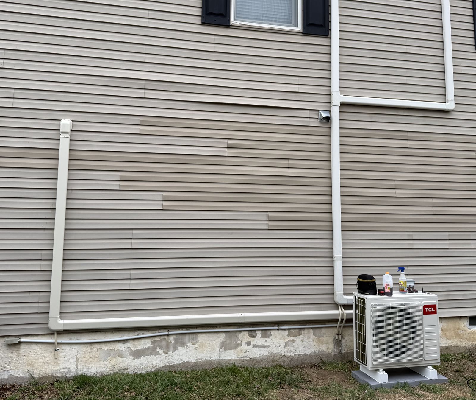 TCL ductless mini-split outdoor unit during installation, with two covered line sets running along a tan-sided house