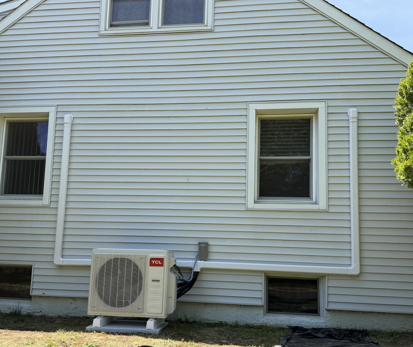TCL ductless mini-split outdoor unit on a pad beside a white-sided house, with white line-set covers along the wall