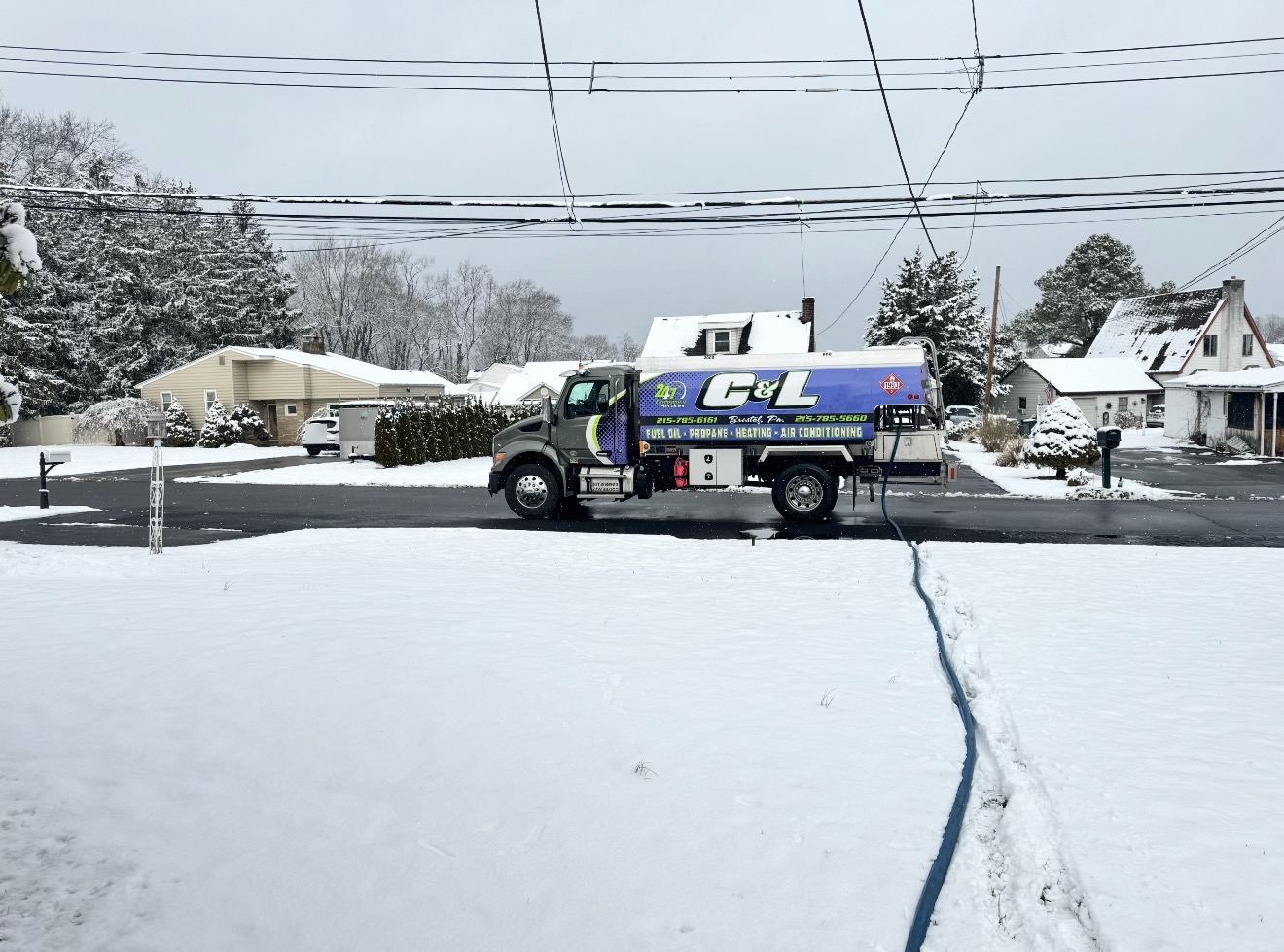 C&L Fuel heating oil delivery truck on a snowy, overcast day, with its delivery hose run across a snow-covered yard