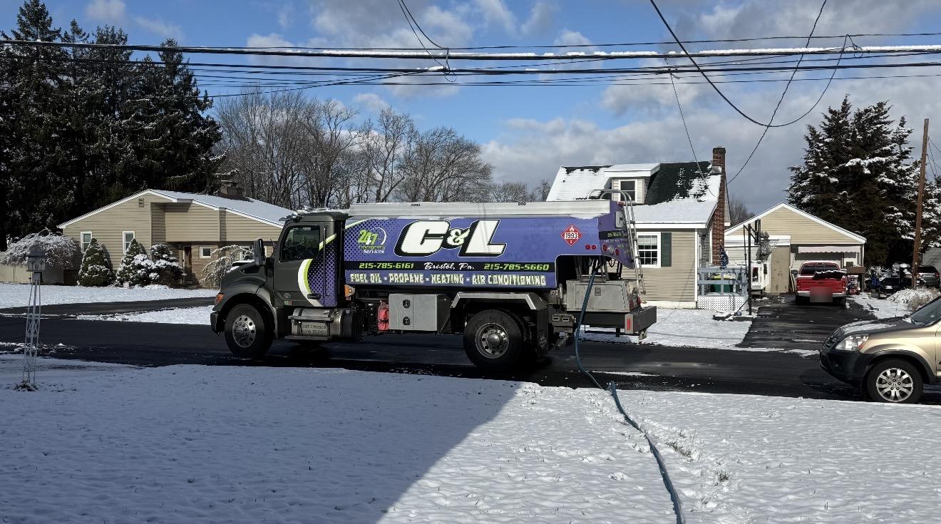 C&L Fuel heating oil delivery truck on a snowy street on a sunny day, with its delivery hose run across the snow