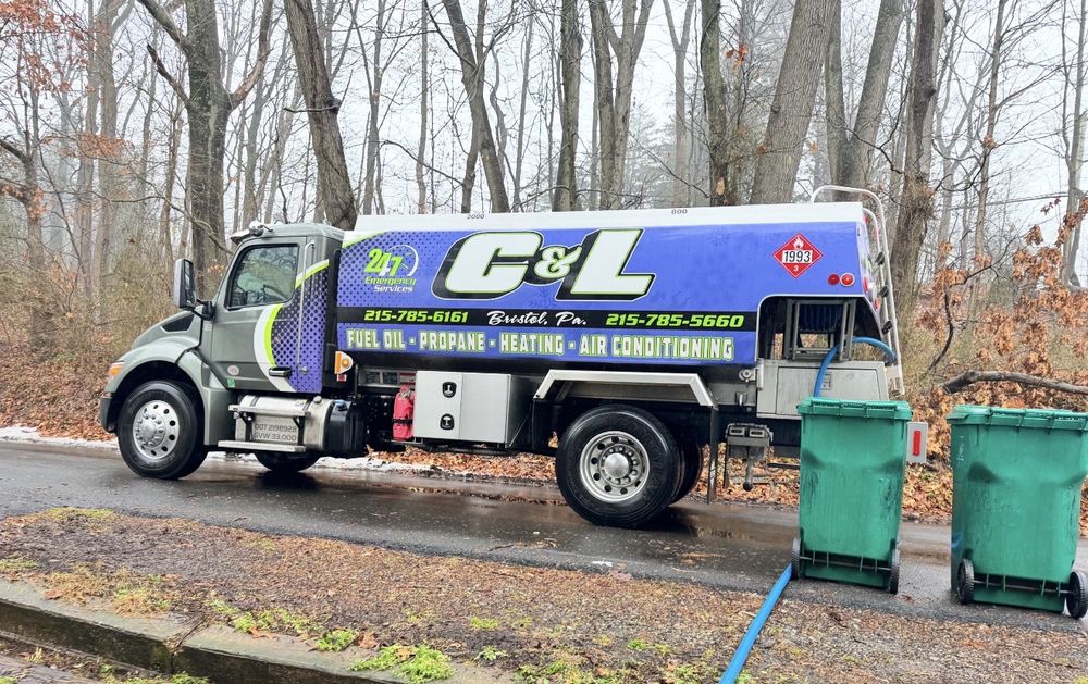 C&L Fuel heating oil delivery truck on a wooded street, with its blue delivery hose run past the trash cans